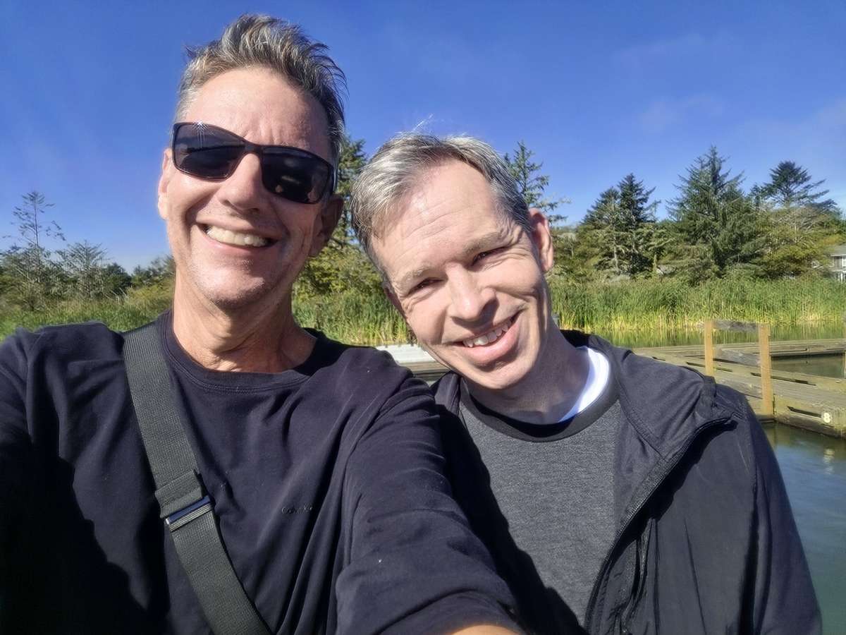 Peter B Dowell and his husband Justin Jones smiling beside the lake.